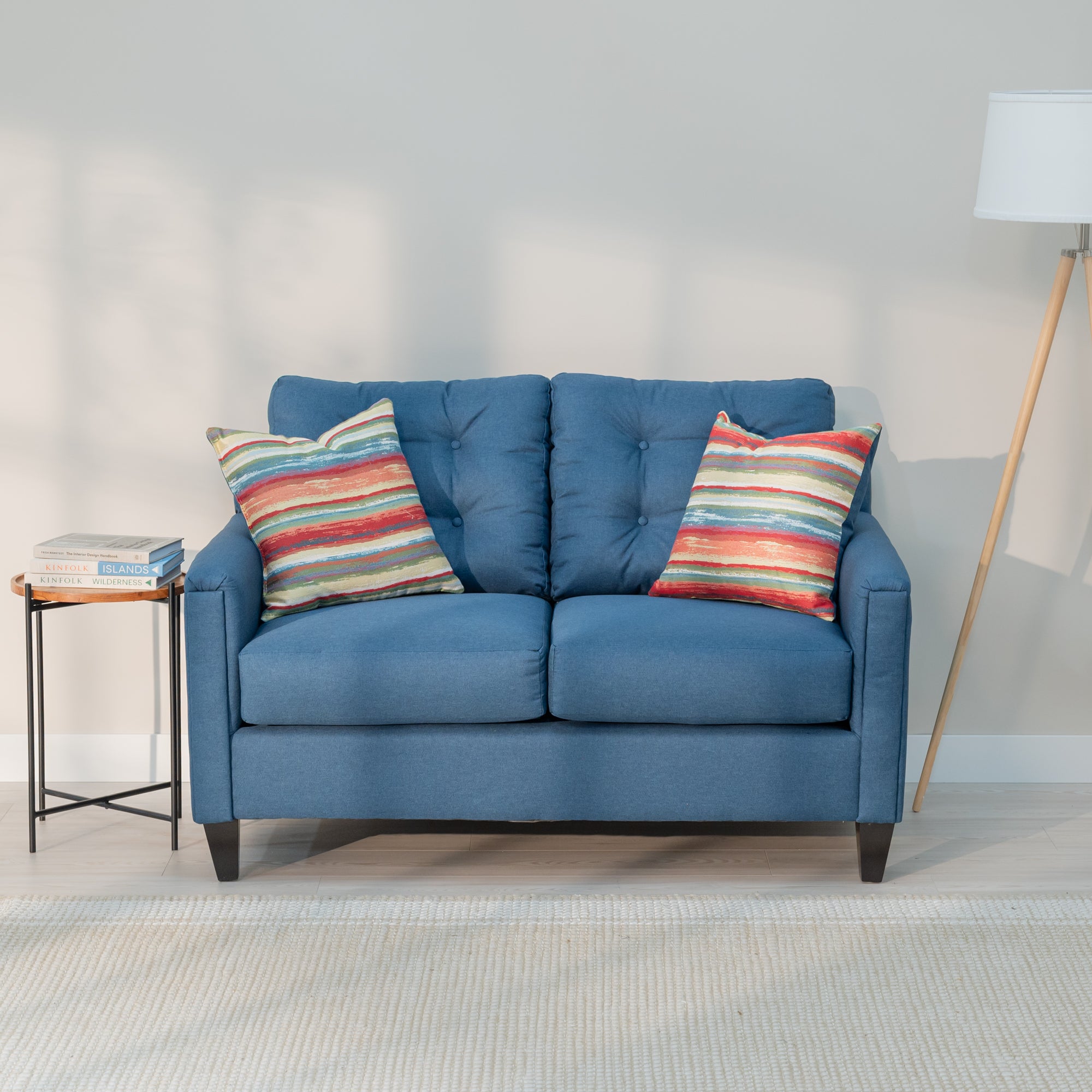 A front view of the denim blue loveseat showing its modern rectangular shape and plush looking tufted back. As well as two throw pillows with rough colorful red, yellow, blue, and green stripes 