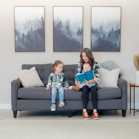 Another view of a beautiful smiling mom reads to a delighted looking toddler and baby while sitting in a modern grey sofa.