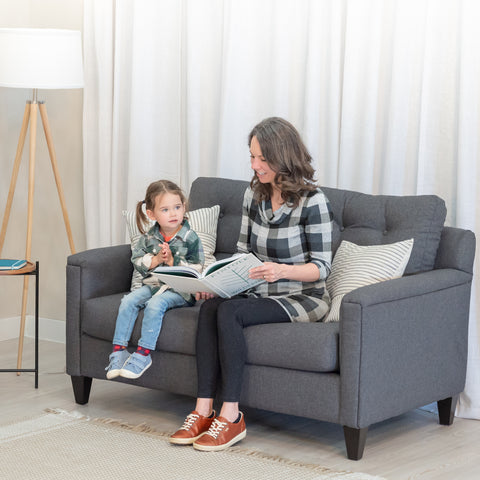 A beautiful smiling mom reads to a delighted looking toddler girl while sitting in a modern grey loveseat chair.