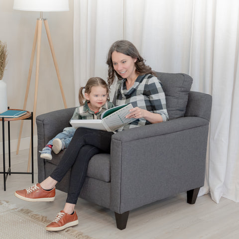 A beautiful smiling mom reads to a delighted looking toddler girl while sitting in a modern grey accent chair. 