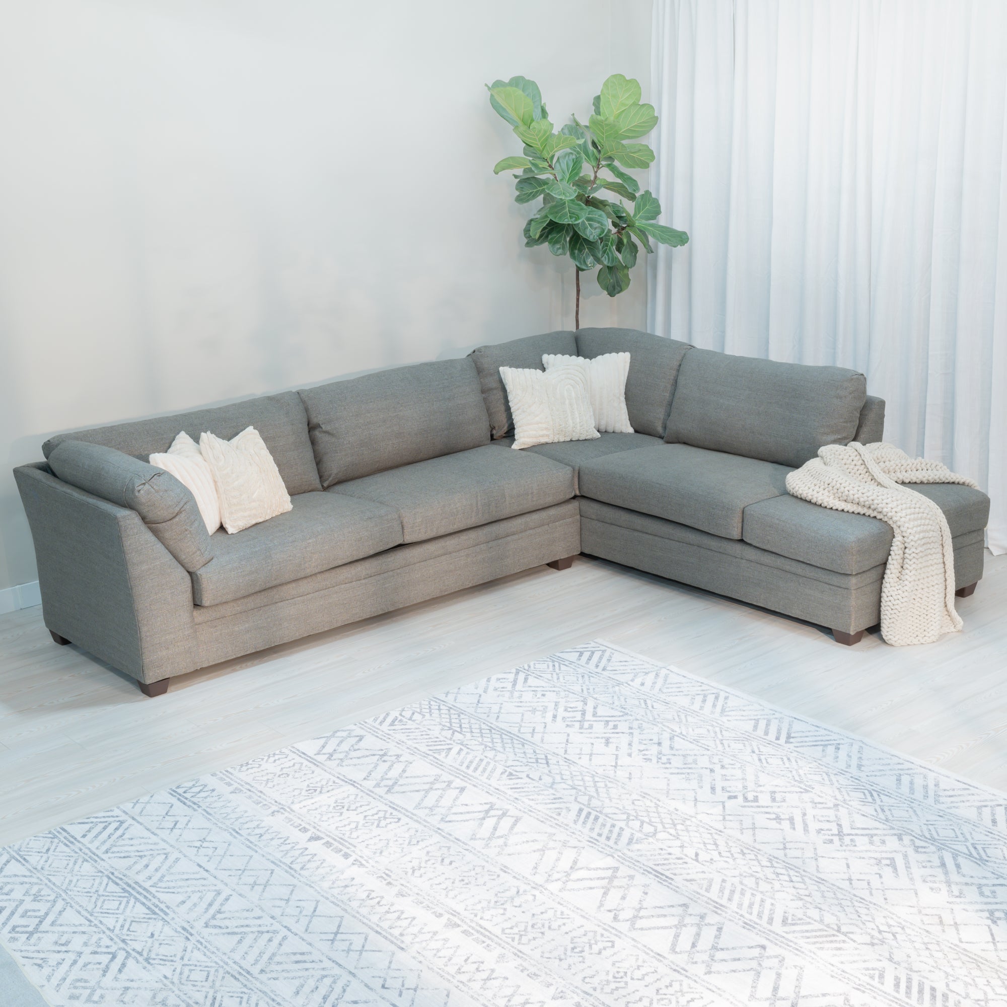 A large light grey sectional sofa with through pillows. The sofa has a backrest style arm on one end and a chase on the other end of the sectional. the back cushions are a plump pillow style cushion and the seat cushions are rectangular with a more structured appearance. the upholstery fabric is a light grey with a  lined style weave and texture. it has thick brown tapered rectangular legs and fluffy faux fur throw pillows. 