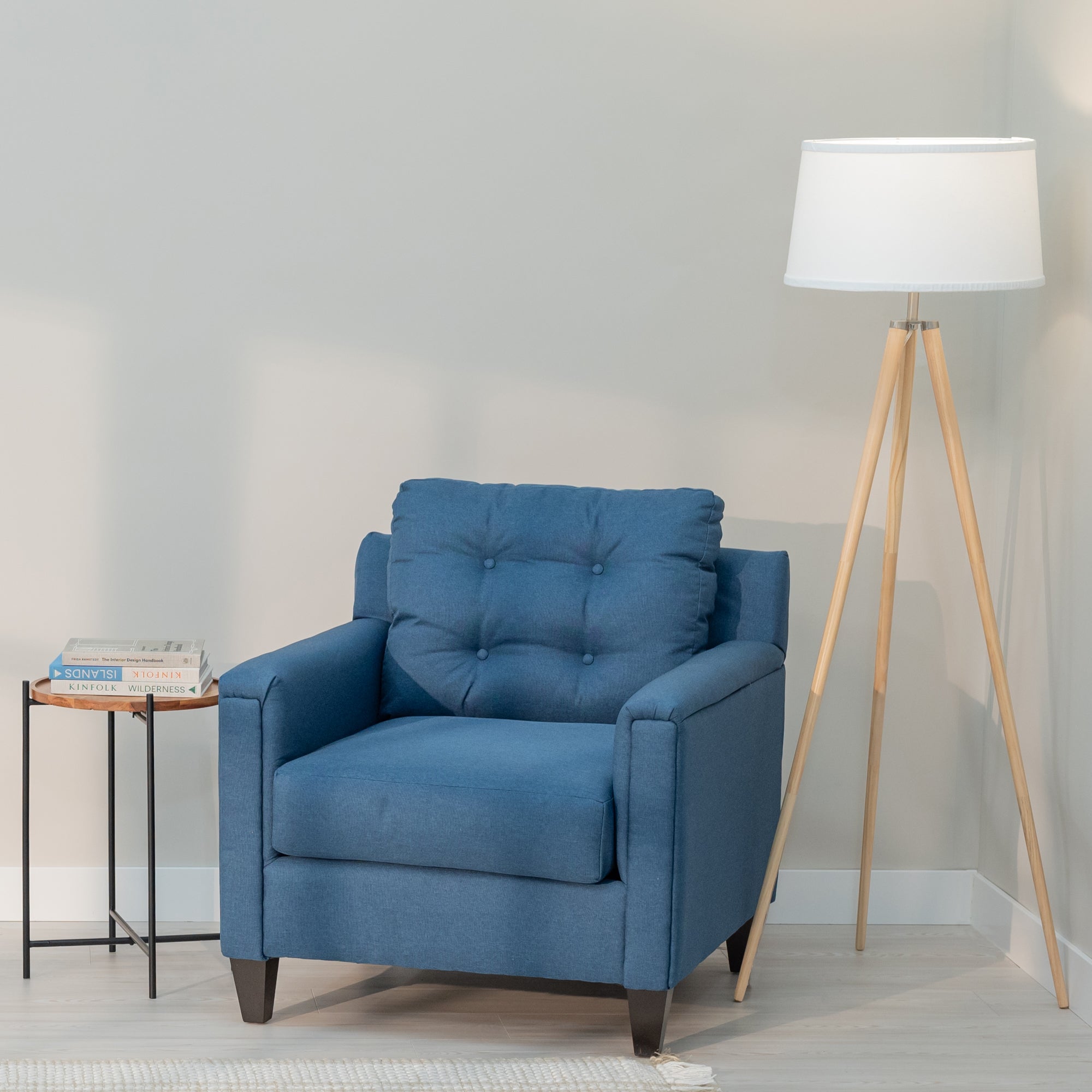 A denim blue accent chair with clean modern lines and a tufted rectangular  bag sits in a generic living room with a side table and floor lamp. The upholstery fabric is blue with lighter colored threads like a denim weave. the chair has tapered dark brown legs .