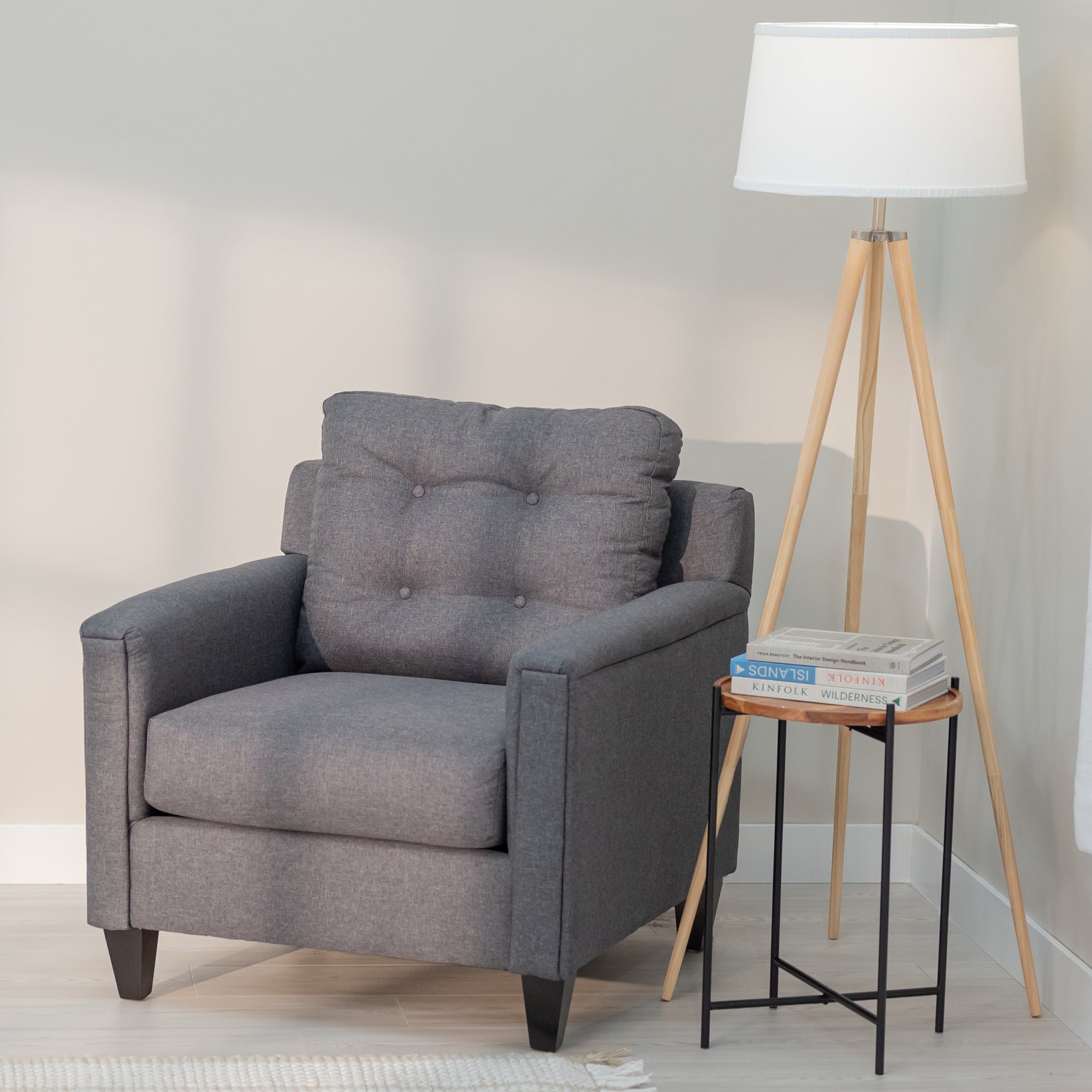 A grey accent chair with clean modern lines and a tufted rectangular  bag sits in a generic living room with a side table and floor lamp. The upholstery fabric is grey with lighter colored threads like a denim weave. the chair has tapered dark brown legs .