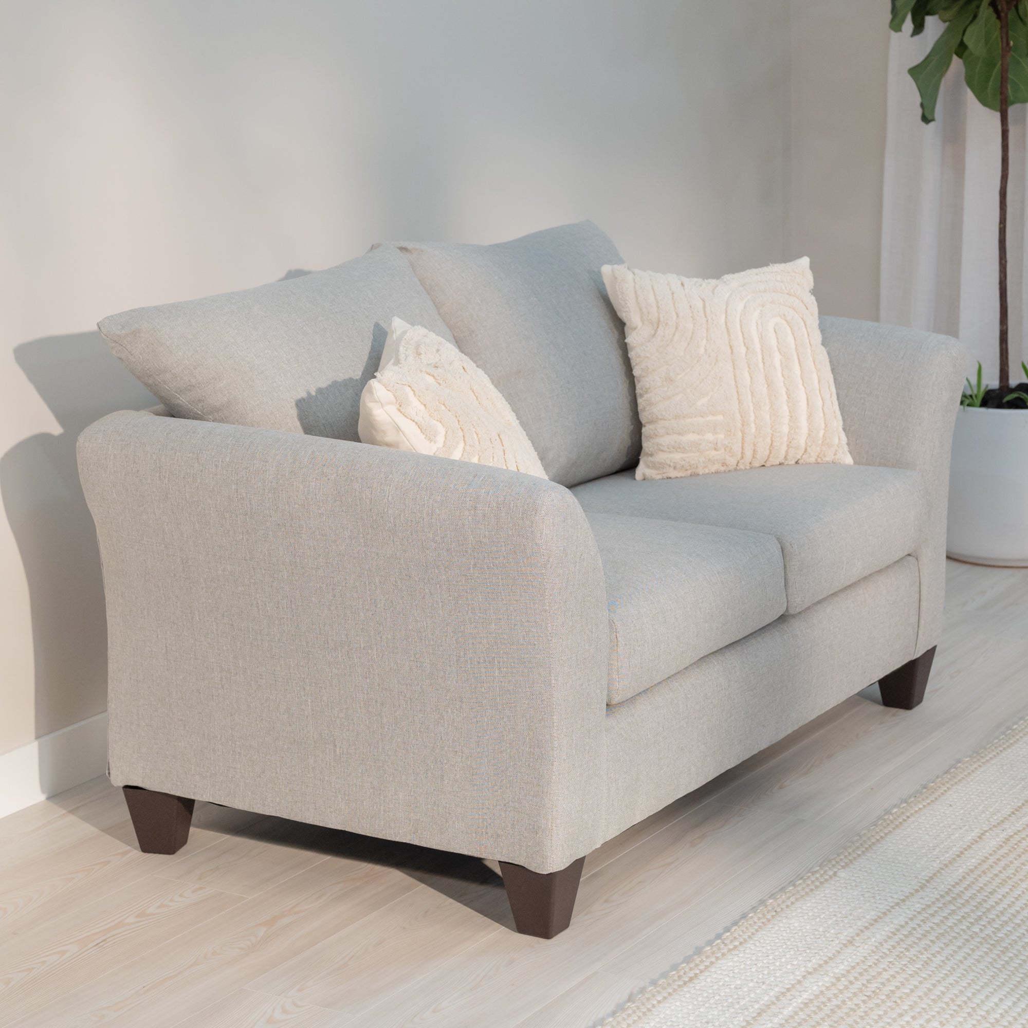 angled view of the loveseat, in a living room Showing the arms curving out from the loveseat like water breaking on the bow of the ship, the plush knife edge pillow style back cushions and plump rectangular seat cushions. All being supported by thick tampered rectangular legs. the loveseat is a light grey with lighter colored grey threads woven through out reminiscent of a linen cloth. The loveseat is decorated with the included throw pillows which a geometric triangular pattern consisting of red, orange, b