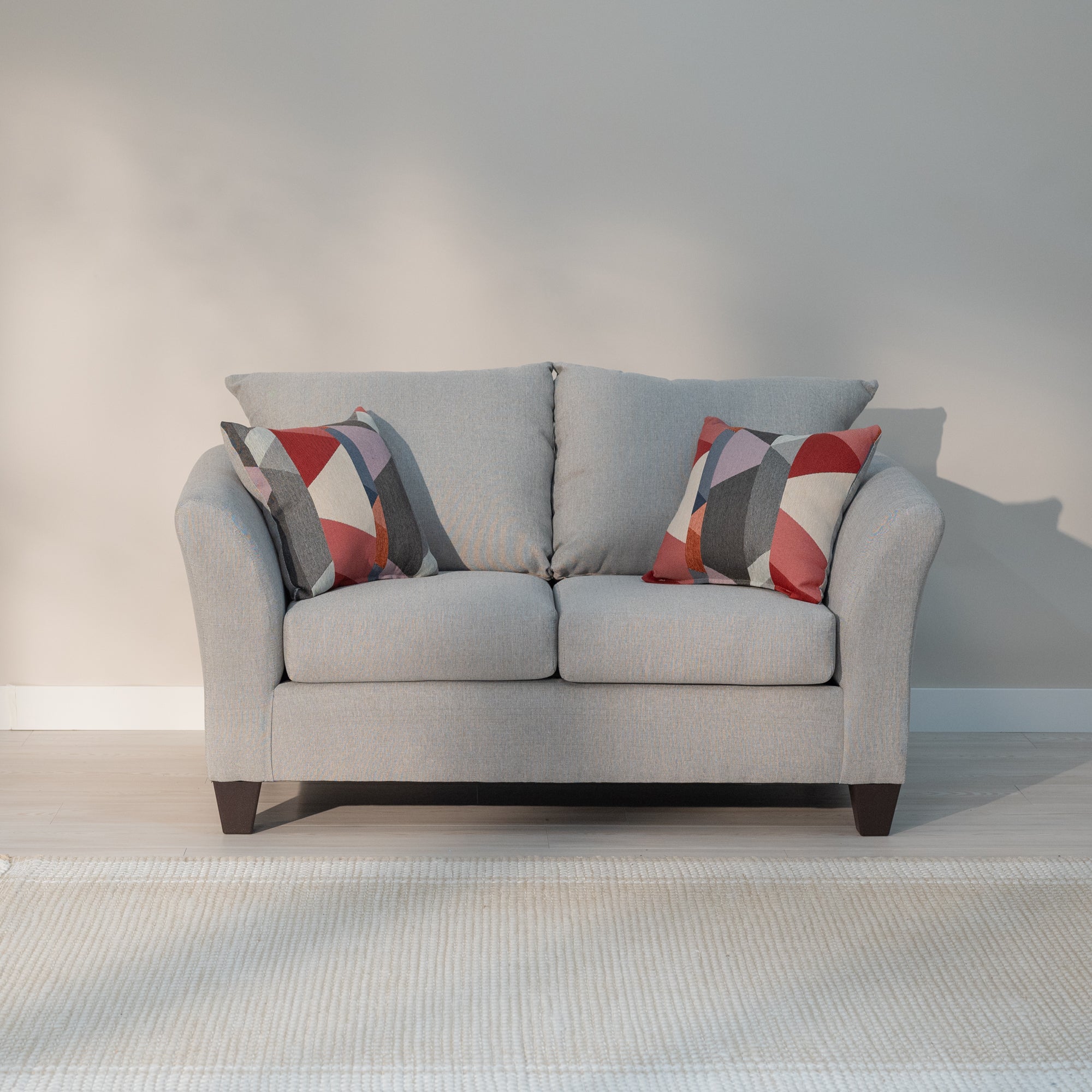 Front view of the loveseat, Showing the arms curving out from the loveseat like water breaking on the bow of the ship, the plush knife edge pillow style back cushions and plump rectangular seat cushions. All being supported by thick tampered rectangular legs. the loveseat is a light grey with lighter colored grey threads woven through out reminiscent of a linen cloth. The loveseat is decorated with the included throw pillows which a geometric triangular pattern consisting of red, orange, blue, grey and whit