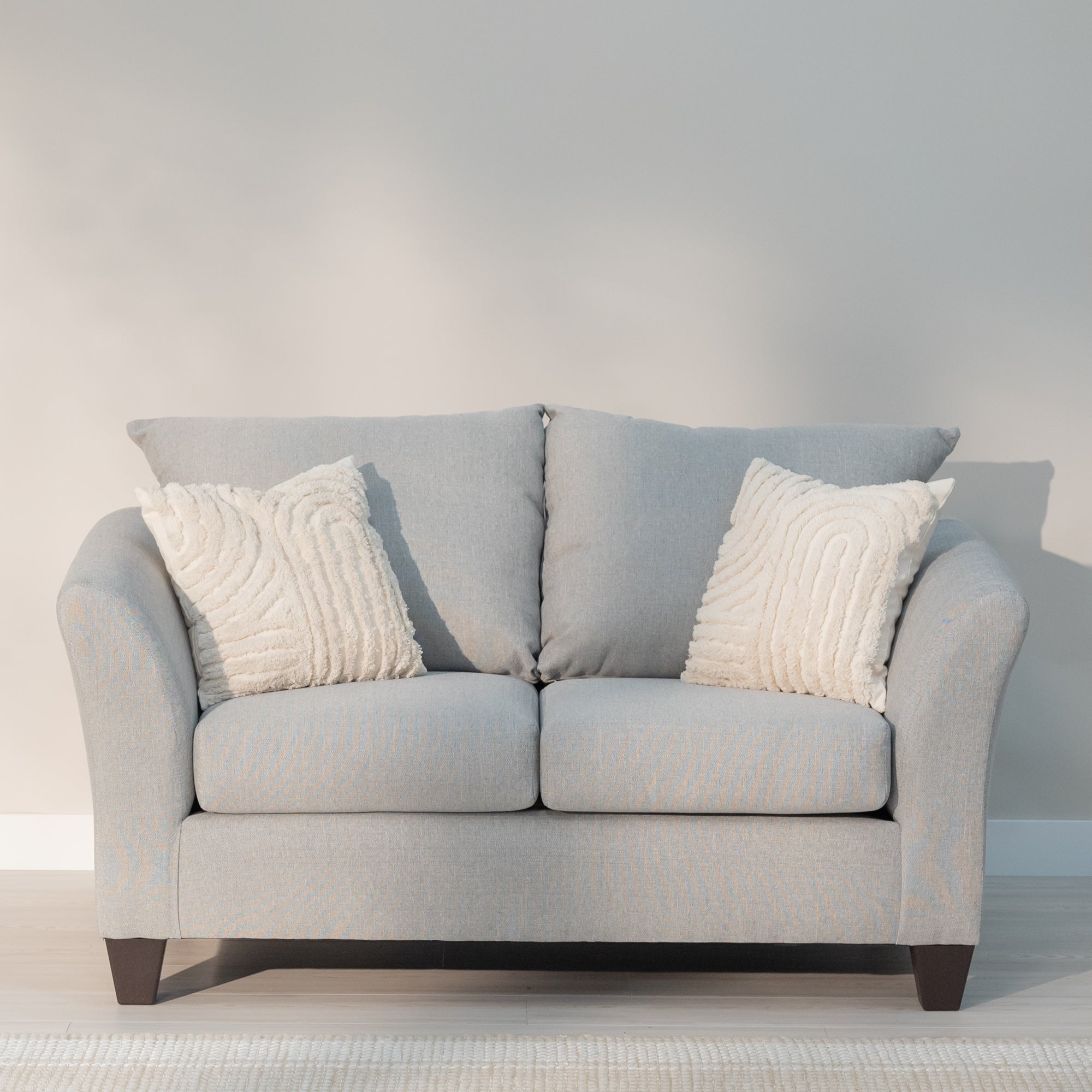 Front view of the loveseat, Showing the arms curving out from the loveseat like water breaking on the bow of the ship, the plush knife edge pillow style back cushions and plump rectangular seat cushions. All being supported by thick tampered rectangular legs. the loveseat is a light grey with lighter colored grey threads woven through out reminiscent of a linen cloth. 