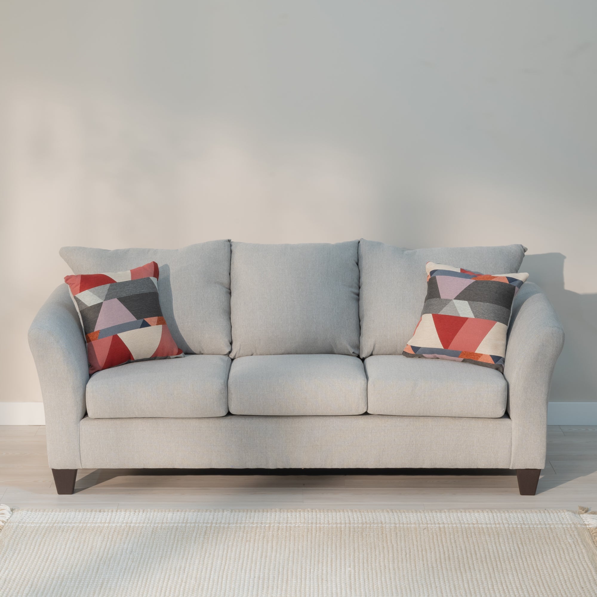 front view of the sofa in a living room, Showing the arms curving out from the loveseat like water breaking on the bow of the ship, the plush knife edge pillow style back cushions and plump rectangular seat cushions. All being supported by thick tampered rectangular legs. the fabric is a light grey with lighter colored grey threads woven through out reminiscent of a linen cloth. The sofa is decorated with the included throw pillows which have a  geometric triangular pattern consisting of red, orange, blue, 