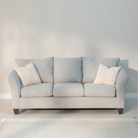 front view of the sofa in a living room, Showing the arms curving out from the loveseat like water breaking on the bow of the ship, the plush knife edge pillow style back cushions and plump rectangular seat cushions. All being supported by thick tampered rectangular legs. the fabric is a light grey with lighter colored grey threads woven through out reminiscent of a linen cloth. The sofa is decorated with two throw pillows