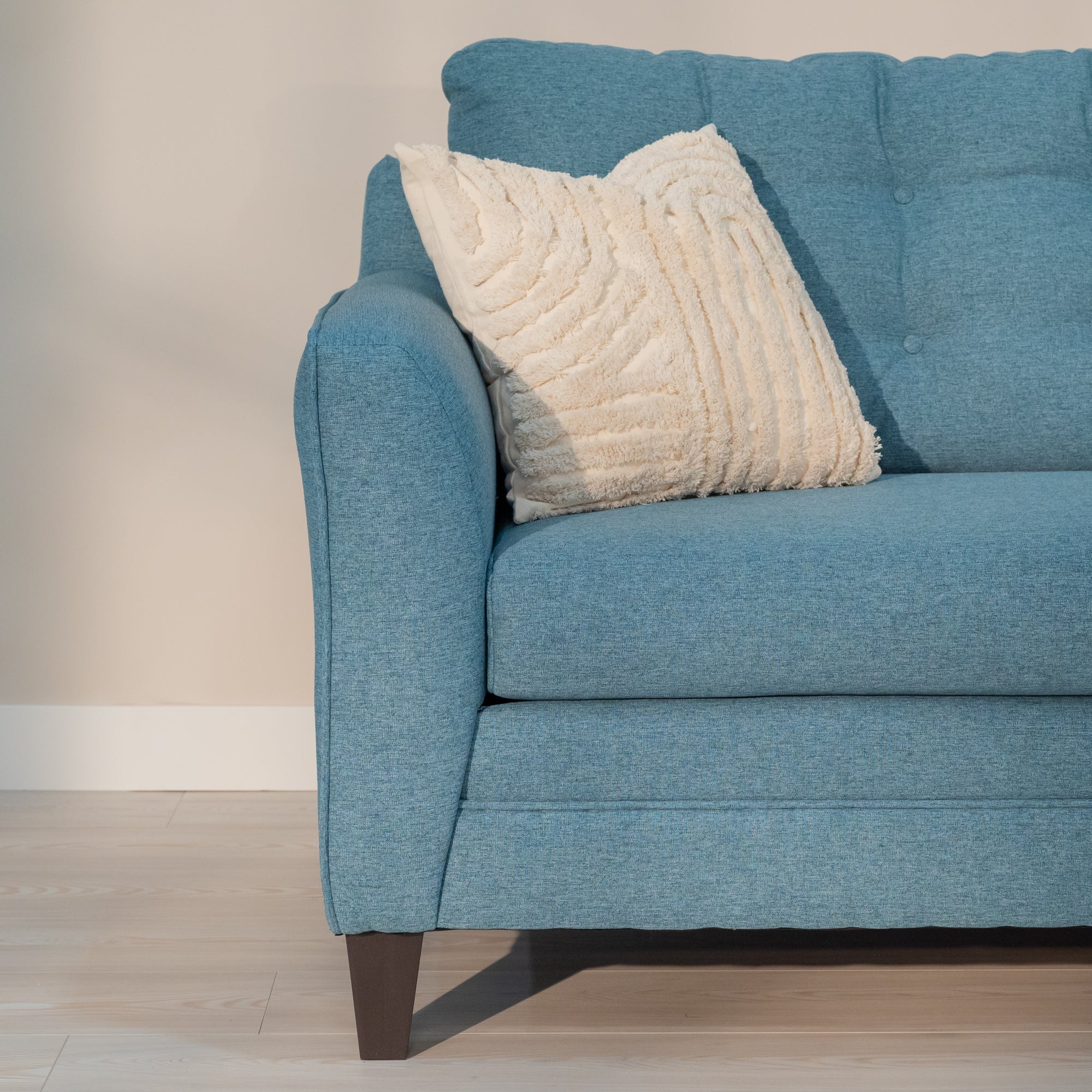 A detailed view of the sofa arm. The outer edge has round piping on the seam while the inside is smooth. the outside edge is straight and the inside edge bows out with padding. The teal fabric has hints of white in it like alien or denim weave, but appears smooth. 