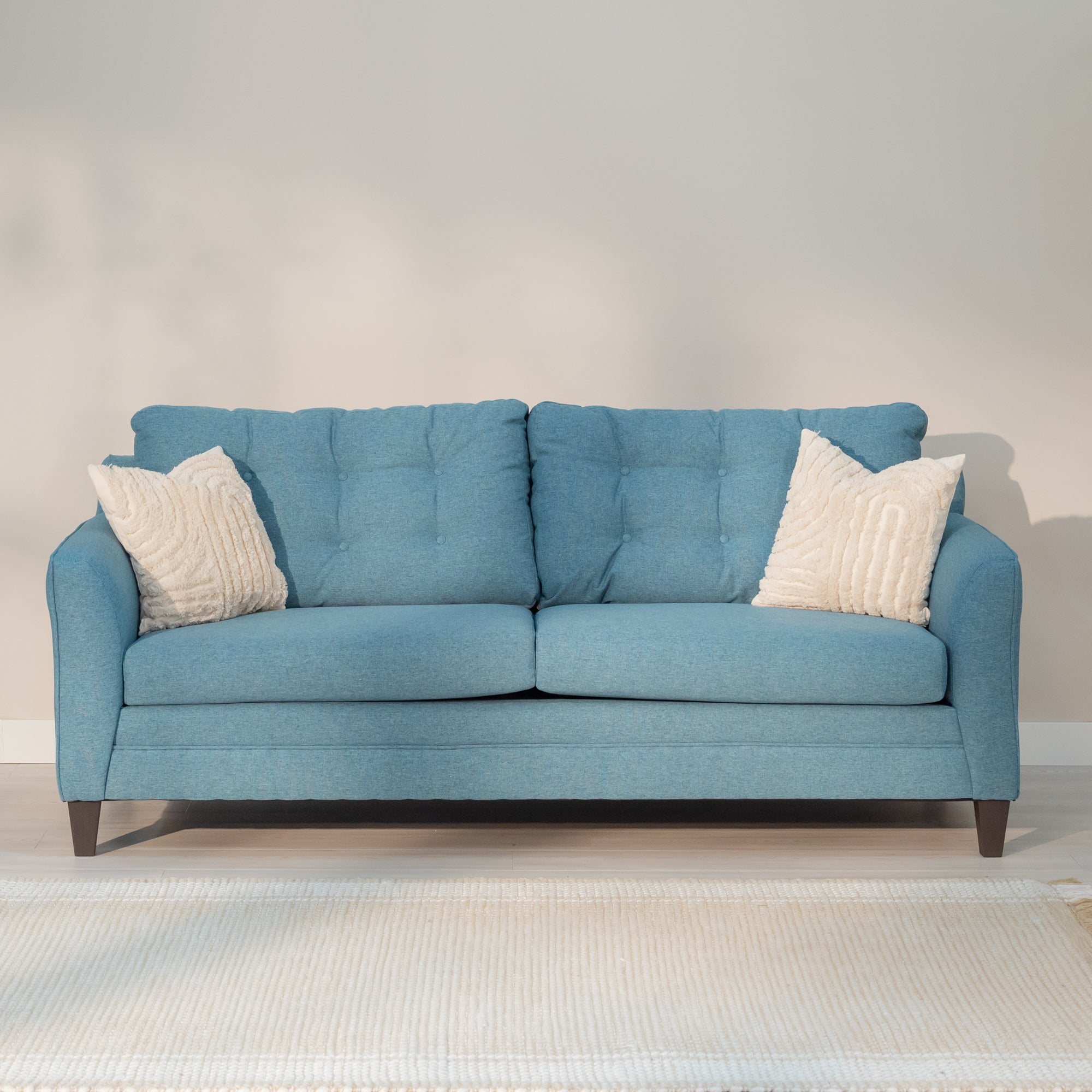 A mid-century style teal colored sofa sits in a living room. It has two wide cushions on the seat and back. the back cushions are tufted and the seat cushions are smooth. the sofa arms are straight ion the our side and curve slightly on the inside.  It sites on tapered dark grey legs about 5 inches tall. 