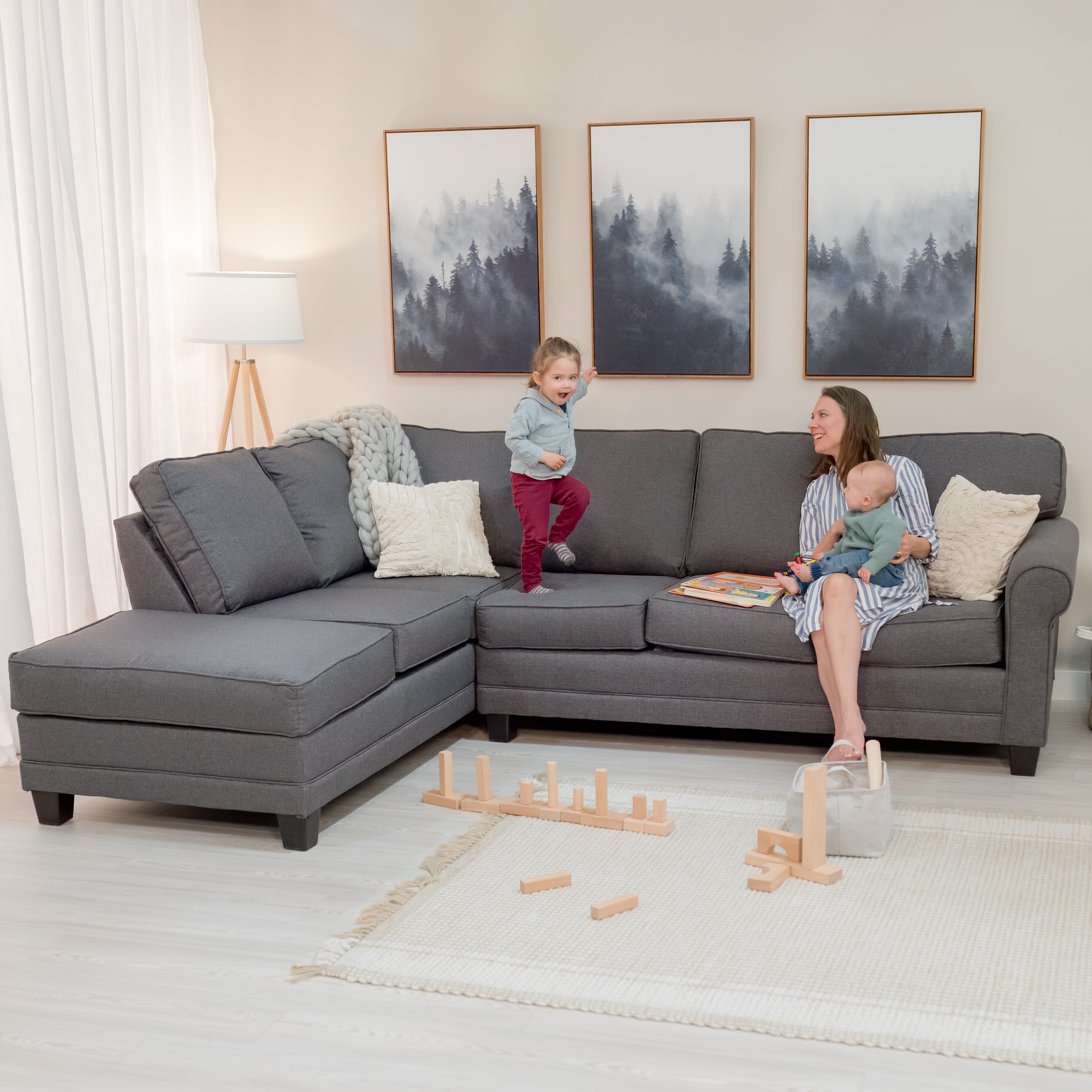 A Pretty mom sits on a large grey sofa sectional with two kids, in a warm and stylish looking living room. One toddler jumps on the sofa while let the baby sits on mom’s lap.  the sofa sectional has a classic rounded arm on the left side and a chase on the right.