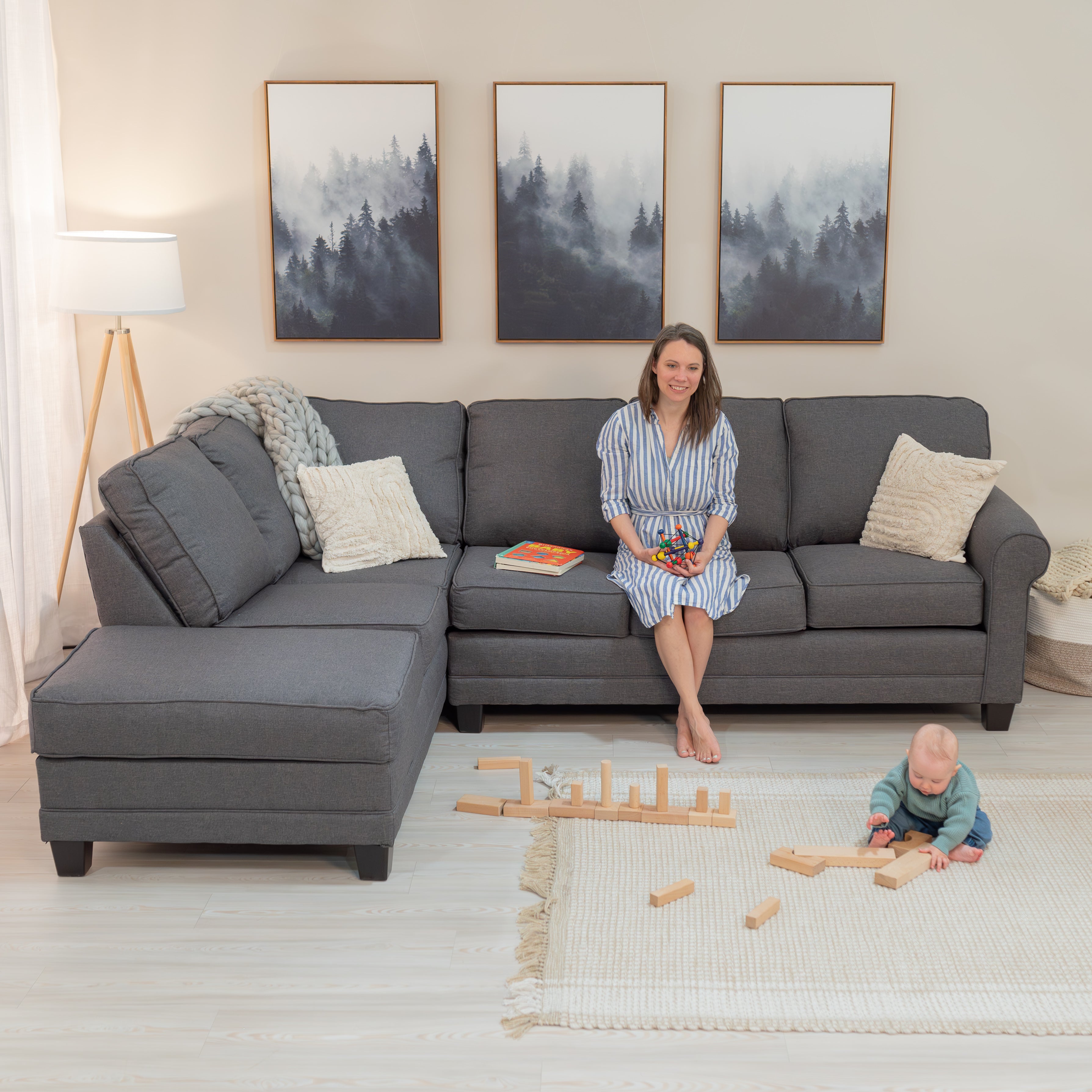 A beautiful mom sits on a large grey sofa sectional holding a baby toy while an adorable baby tries to play with blocks on the floor to be like her big sister. 