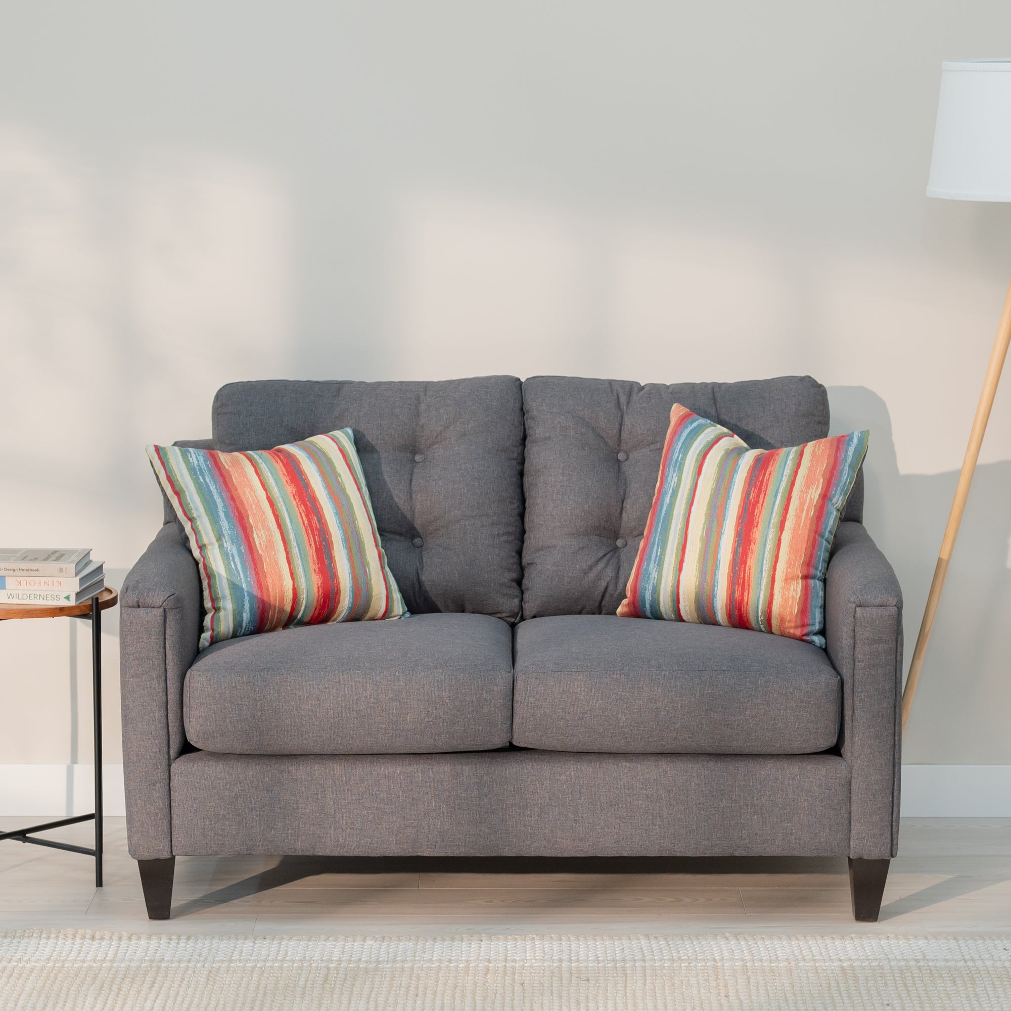 A front view of the grey loveseat showing its modern rectangular shape and plush looking tufted back. As well as two throw pillows with rough colorful red, yellow blue and green stripes 