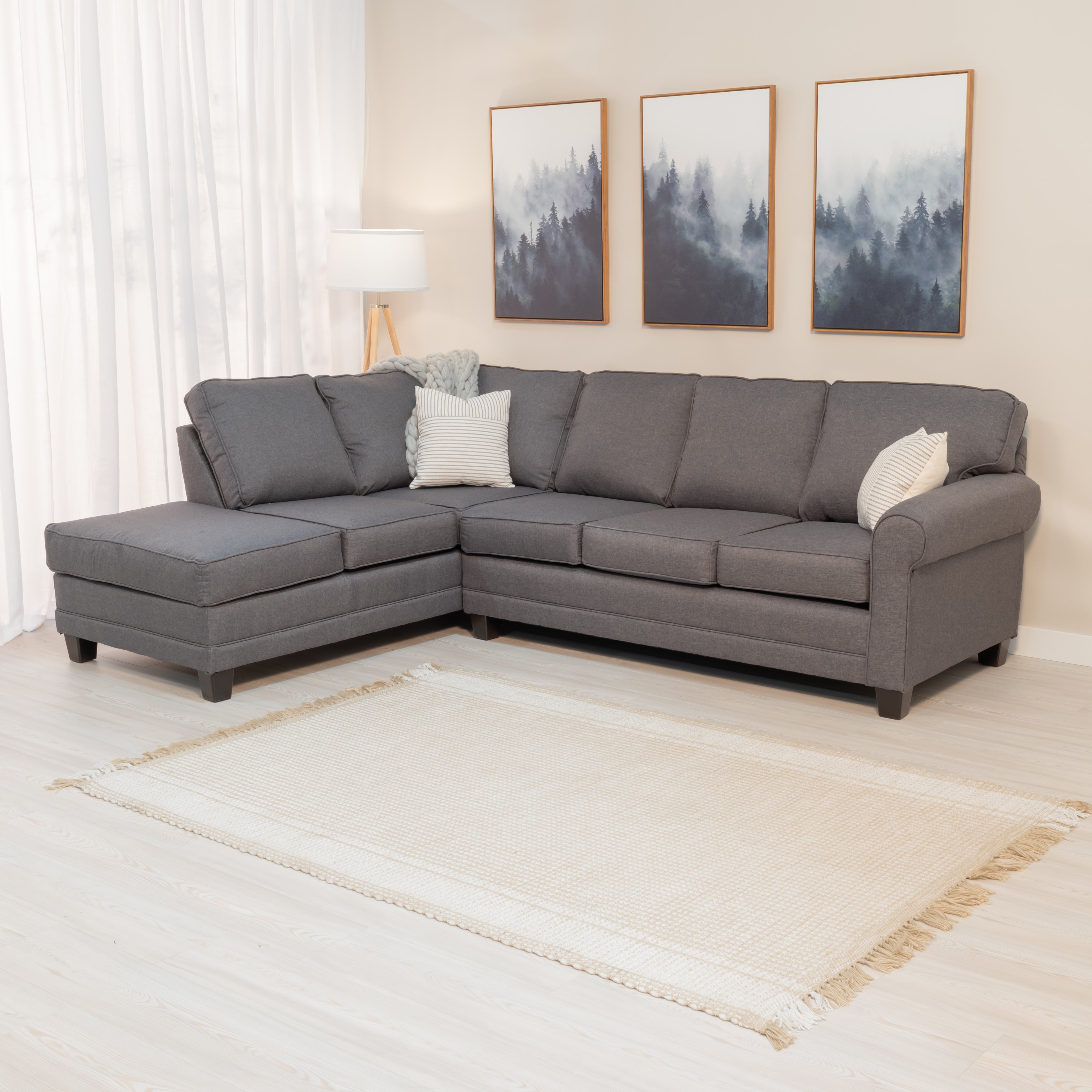 A large grey sectional sofa with throw pillows in a sunny living room. the sofa sectional has a classic rounded arm on the left side and a chase on the right. it has dark grey rectangular legs and and plush looking cushions with round piping on the seams. The upholstery fabric is grey with lighter colored threads like a denim weave.   