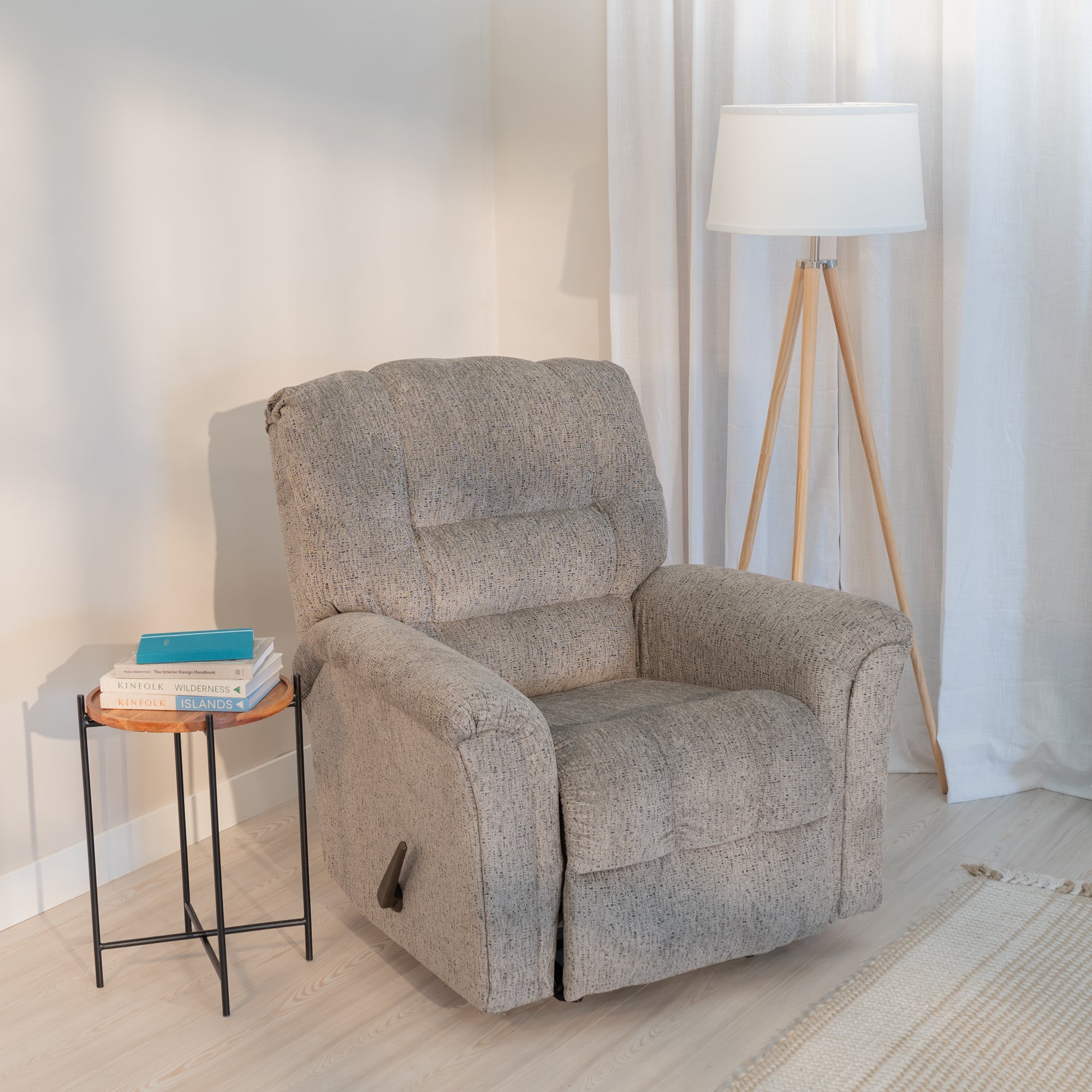 A soft fluffy rocking recliner sitting in a living room corner with a side table and lamp. It is light gray with dark charcoal flecks in the soft upholstery. 