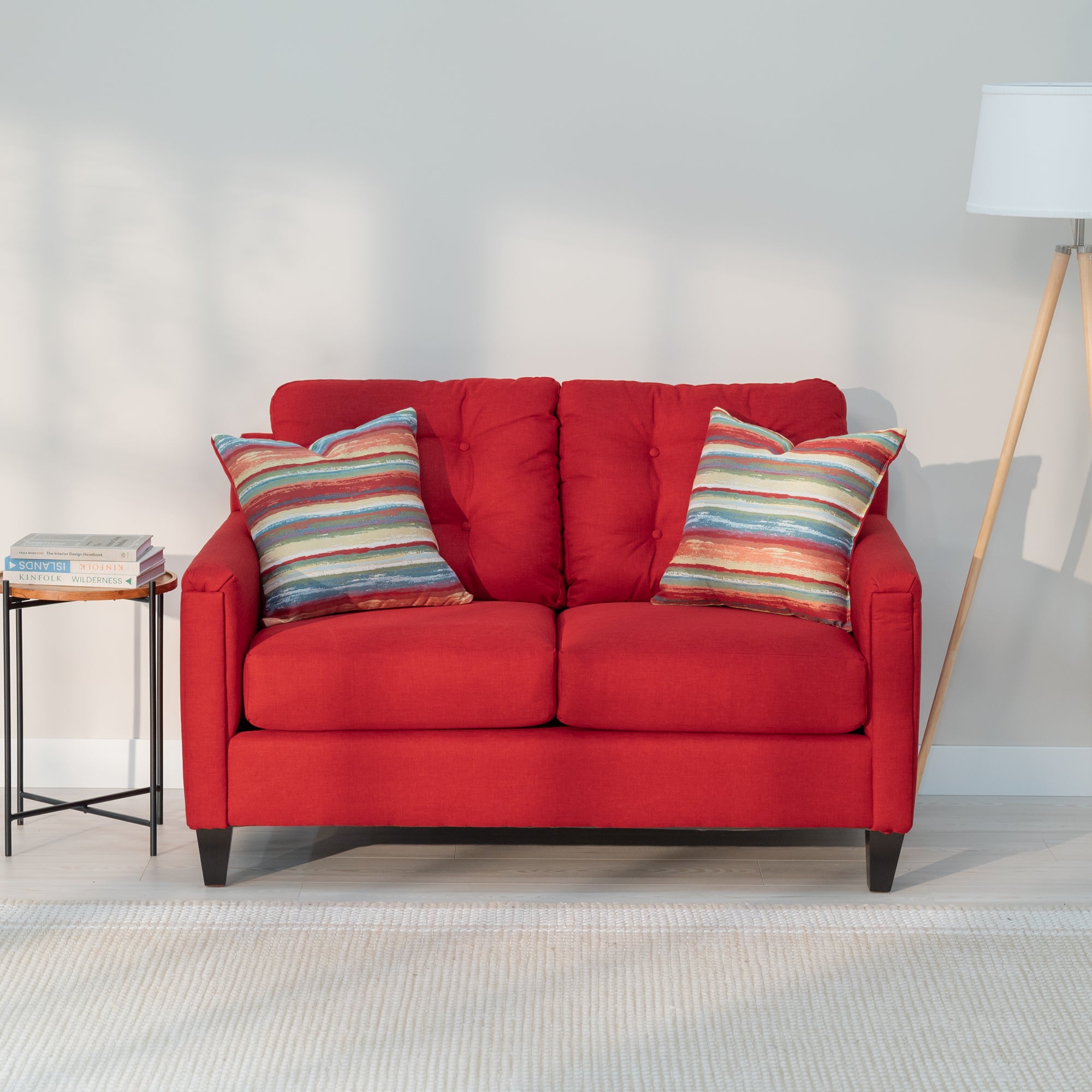 A front view of the red loveseat showing its modern rectangular shape and plush looking tufted back. As well as two throw pillows with rough colorful red, yellow, blue, and green stripes 